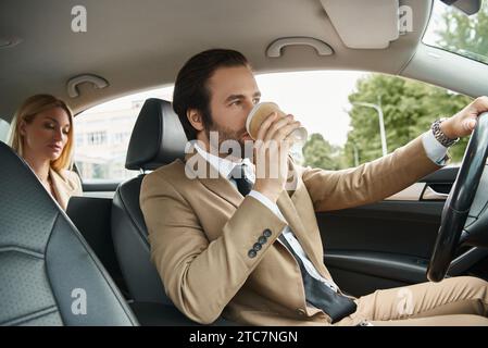 homme élégant dans l'usure formelle buvant du café à et conduisant la voiture avec femme d'affaires blonde sur le siège arrière Banque D'Images