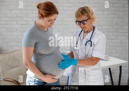 Femme enceinte consultant un médecin. Gynécologue caucasienne âgée tient les mains sur le ventre d'une patiente enceinte. Banque D'Images