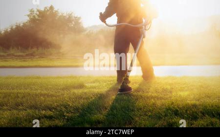 Plan rapproché d'un travailleur municipal tenant une tondeuse à gazon, taillant l'herbe dans un parc public pendant un lever de soleil vibrant. Entretenir la pelouse à l'aube avec Banque D'Images