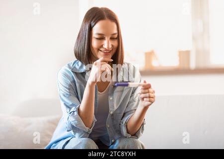 Femme enceinte à la maison tenant joyeusement le résultat positif du test de grossesse Banque D'Images