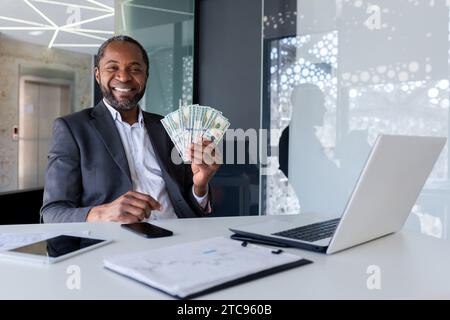 Portrait rapproché d'un homme d'affaires afro-américain assis dans le bureau à un bureau, tenant un fan d'argent, pointant et souriant à la caméra. Banque D'Images