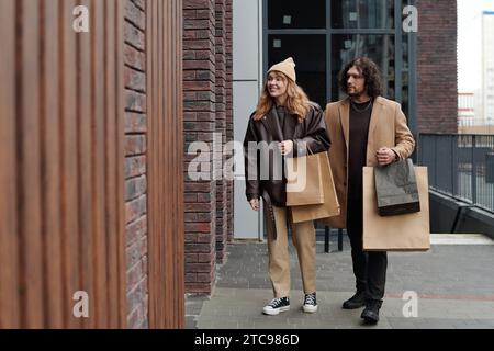 Les jeunes acheteurs en vêtements décontractés élégants portant des sacs de papier avec des achats tout en marchant le long des vitrines du Black Friday Banque D'Images