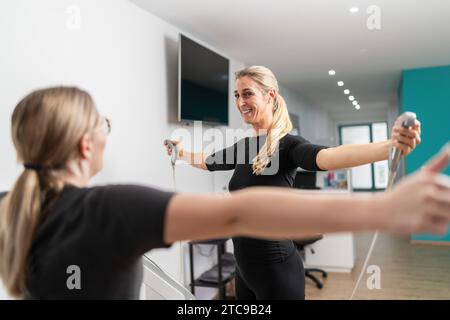 Le client de fitness souriant tout en tenant l'analyseur d'échelle de composition corporelle gère le test interne, étendant les bras vers l'entraîneur dans un EMS - Studio. Banque D'Images