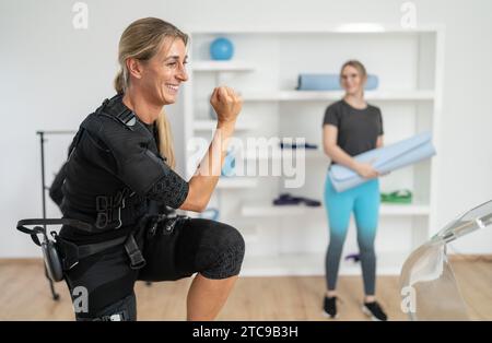 Femme en costume EMS faisant des squats avec entraîneur tenant tapis de yoga pour le prochain exercice d'entraînement dans un studio EMS Banque D'Images