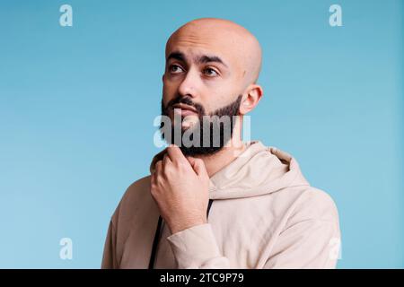 Homme arabe regardant loin avec l'expression du visage pensive et frottant le menton pensivement. Jeune barbu chauve pensant et prenant des décisions tout en posant avec un geste perplexe Banque D'Images