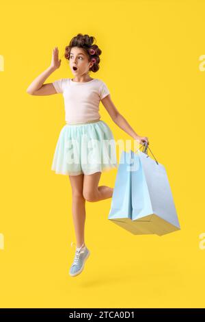 Petite fille choquée avec des bigoudis et des sacs à provisions sautant sur fond jaune Banque D'Images