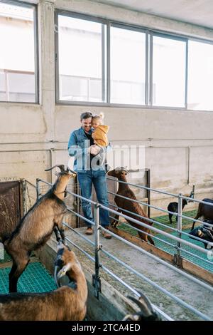 Père avec une petite fille dans les bras se tient près d'un paddock avec une chèvre appuyée sur la clôture Banque D'Images