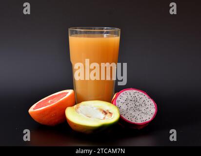 Un grand verre d'un mélange de jus de fruits tropicaux sur fond noir, à côté d'une demi-mangue mûre, pitaya et pamplemousse. Gros plan. Banque D'Images