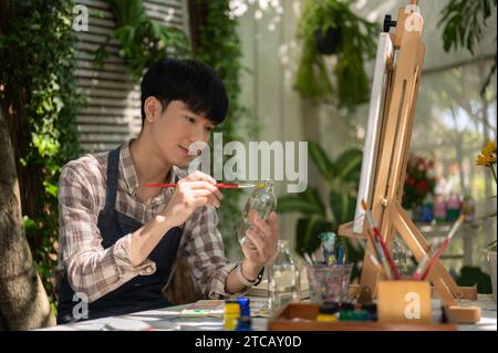 Un jeune homme asiatique positif et beau peint et colore une bouteille en verre avec une couleur acrylique dans son jardin à la maison, fabriquant à la main une bouteille décorative Banque D'Images