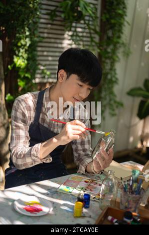Un jeune homme asiatique positif et beau peint et colore une bouteille en verre avec une couleur acrylique dans son jardin à la maison, fabriquant à la main une bouteille décorative Banque D'Images