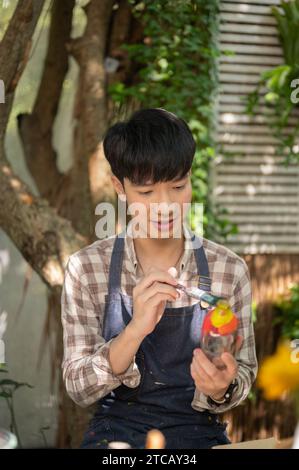 Un jeune homme asiatique positif dans un tablier rejoignant un atelier d'art, peignant une bouteille en verre, fabriquant à la main une bouteille décorative. Concept de passe-temps Banque D'Images