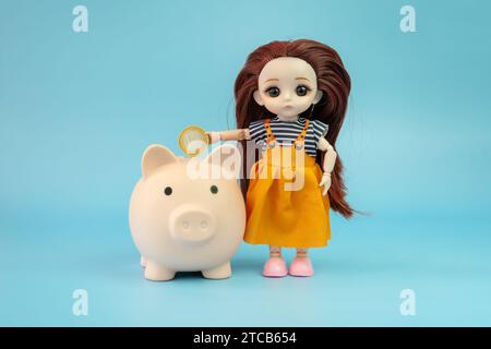 Une petite poupée enfant avec une pièce dans les mains sur un fond bleu. La poupée met une pièce dans la tirelire. Banque D'Images