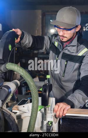 Jeune homme brunette portant une casquette dans une veste grise par profession un charpentier coupe des planches de bois avec une scie circulaire sur une table d'établi en A. Banque D'Images