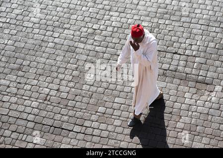 Marrakech, Maroc : un vieil homme marocain portant un caftan blanc et une casquette kufi rouge se promène sur la place Jemaa el Fnaa, Médina de Marrakech. Banque D'Images