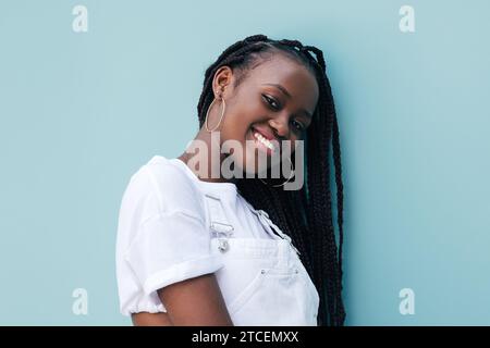 Portrait de jeune femme souriante avec des tresses appuyées sur le mur bleu regardant la caméra Banque D'Images