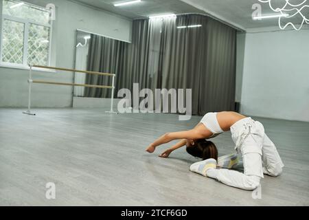 femme afro-américaine flexible en vêtements de sport blancs répétant la danse moderne dans un studio spacieux Banque D'Images