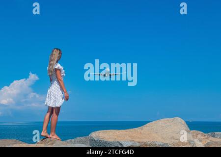 Seaside Serendipity : fille en robe blanche, pierres, mer bleue et un avion Banque D'Images