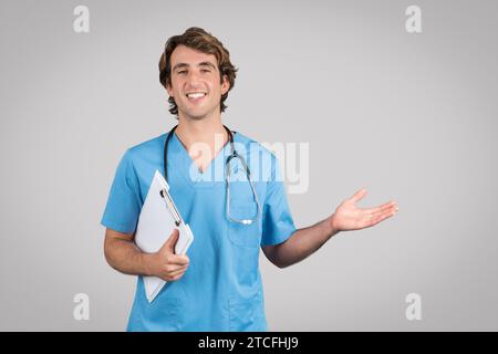 Médecin masculin excité dans Scrubs tenant le presse-papiers et montrant quelque chose sur la paume vide pointant avec la main à l'espace de copie Banque D'Images