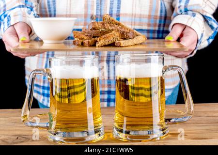 Deux tasses en verre avec de la bière sur le fond d'une fille avec des collations à la bière sur une planche de service. Accent sélectif sur les tasses avec de la bière. Banque D'Images