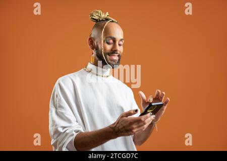 Homme gay Latino avec maquillage lumineux utilisez smartphone isolé fond orange. Confiant brésilien gay habillé blanc à l'aide du téléphone portable, l'envoi de message dans les médias sociaux ou faire des achats en ligne Banque D'Images