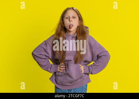 Joyeux drôle jeune fille d'école rousse montrant la langue faisant des visages à la caméra, se trompant, plaisantant, apantant avec un visage idiot, taquinant, intimidant, abusant. Enfant féminin préadolescent sur fond jaune Banque D'Images