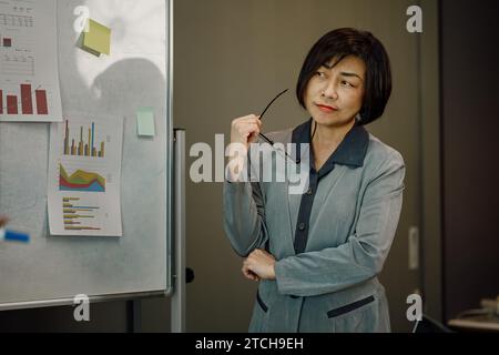 Femme d'affaires senior sérieuse analysant les statistiques de ventes sur tableau blanc. Banque D'Images