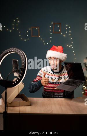 Homme arabe influenceur dans Santa Hat déballant l'ordinateur portable et enregistrant la vidéo, assis devant le téléphone portable, partageant les achats de magasin d'Internet Banque D'Images