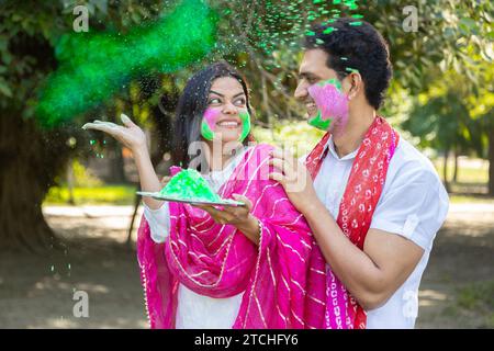 Heureux jeune couple indien portant kurta blanc jetant, mettant de la couleur sur le visage. Célébrer Holi ensemble en plein air dans le parc ou le jardin. Banque D'Images