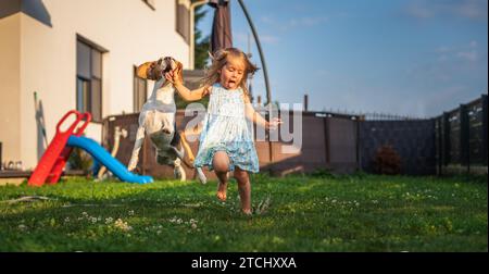 Bébé fille courant avec le chien beagle dans le jardin le jour d'été. Concept d'animal domestique avec enfants. Chien chassant l'enfant avec une balle de tennis Banque D'Images