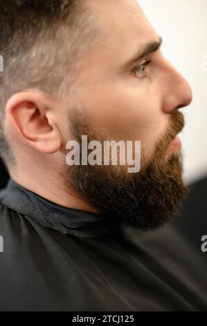 Un jeune homme avec une barbe dans une cape noire est assis sur une chaise dans un salon de coiffure. Le client attend une coupe de cheveux et le rasage de la barbe. Banque D'Images