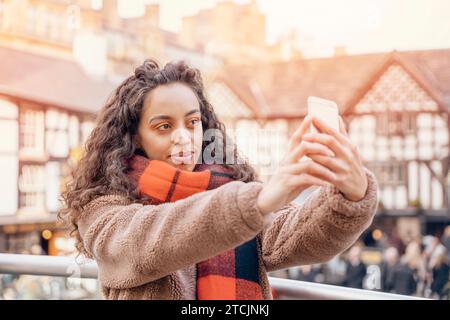 Une femme gaie qui prend des selfies sur un smartphone Une dame brune dans une veste et une écharpe orange souriant et voyageant en Europe. Banque D'Images
