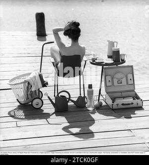 style de vie des années 1950. Une jeune femme au soleil portant un bikini. Dans l'image, il y a beaucoup d'objets opportuns en plastique qui étaient populaires à l'époque. Surtout lorsque vous êtes en vacances d'été ou en camping. Un ensemble de tasses et soucoupes sur la table. Un cas pratique pour les transporter. 1958 Kristoffersson réf DB95-12 Banque D'Images