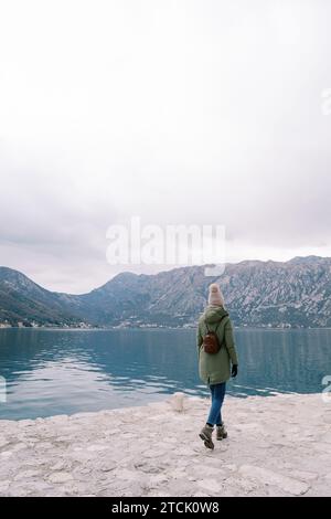 Femme avec un sac à dos marche le long de la jetée par la mer et regarde les montagnes. Vue arrière Banque D'Images