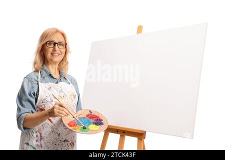 Artiste féminine debout à côté d'une toile de peinture et tenant un pinceau et une palette isolé sur fond blanc Banque D'Images