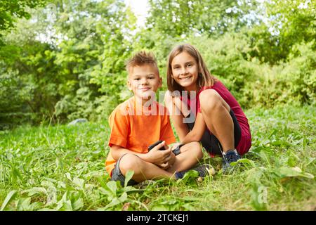 Portrait d'une fille souriante accroupie à côté d'un garçon assis avec un téléphone intelligent au parc Banque D'Images