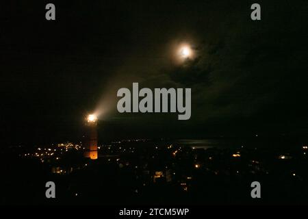 Phare de nuit, lumières dans le village ci-dessous, envoyant un faisceau de lumière dans la direction de la lune Banque D'Images