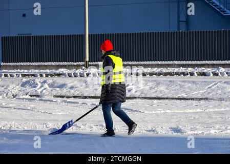 Femme dégage la route de la neige avec une pelle. Une femme au travail portant un gilet jaune. Nettoyage de la zone. Banque D'Images