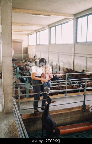 Maman embrasse une petite fille assise sur la clôture du paddock et regarde la chèvre Banque D'Images