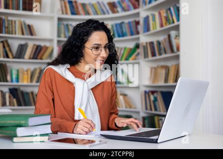 Étudiante satisfaite écoute une conférence en ligne sur un ordinateur portable, écrit quelque chose dans un cahier, s'assoit à une table dans un bureau, apprentissage à distance. Banque D'Images