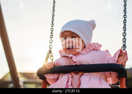 Adorable baby girl avec de grands beaux yeux et un bonnet s'amusant sur une balançoire ride à une aire de jeux dans un parc ensoleillé Banque D'Images