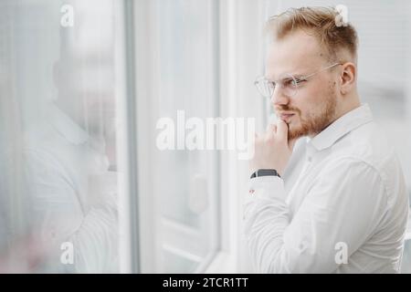 Un jeune homme d'affaires dans une chemise blanche regarde soigneusement par la fenêtre d'un gratte-ciel Banque D'Images