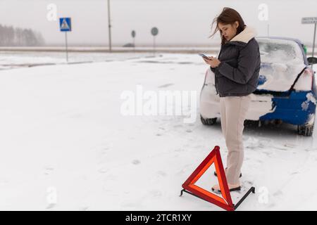 Une jeune femme qui a écrasé sa voiture sur une route glissante d'hiver dans la neige appelle une dépanneuse avec un triangle d'urgence à côté d'elle près de la voiture Banque D'Images