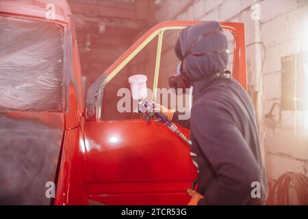 Réparation de carrosserie d'une camionnette, peinture la carrosserie en rouge, un mécanicien dans un respirateur peint une camionnette à l'aide d'un pistolet pulvérisateur Banque D'Images