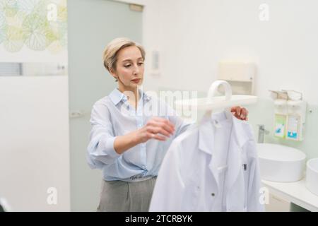 Le médecin professionnel d'âge moyen entre dans le bureau, enlève la robe du porte-manteau et le met elle-même. Le médecin aux cheveux gris se prépare Banque D'Images