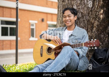 Jeune mâle asiatique positif se repose sous l'arbre dans un parc avec sa guitare acoustique, en pratiquant la guitare. Concept de loisirs et de hobby Banque D'Images