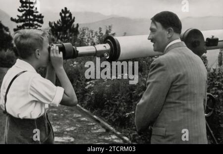 Adolf Hitler est représenté accompagnant un enfant qui regarde à travers un télescope. Cette image, capturant un moment apparemment intime et personnel, a probablement été utilisée comme un outil de propagande pour dépeindre Hitler sous un jour plus relatable et compatissant. La juxtaposition d'un dirigeant puissant avec un enfant dans un scénario d'apprentissage ou de découverte était une tactique pour adoucir son image publique et attirer la population en général, mettant en valeur un aspect différent de son personnage. Banque D'Images