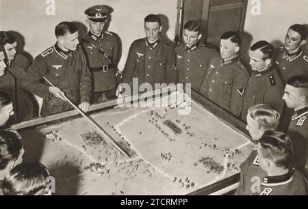 Les recrues allemandes sont photographiées rassemblées autour d'une « table de sable », un outil traditionnel utilisé dans l'entraînement militaire pour enseigner la tactique et la stratégie. Ce modèle interactif leur permet de visualiser et de comprendre divers scénarios et stratégies de champ de bataille. Banque D'Images