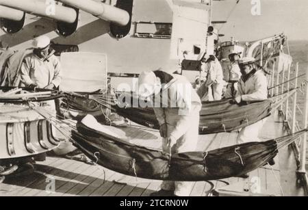 Sur cette image, des Marines allemands de la Kriegsmarine sont vus accrochant leurs hamacs sur le pont d'un cuirassé, une routine nécessaire pour les aérer. Compte tenu de l'espace limité et de la nature de leurs couchages à bord du navire, l'aération régulière des hamacs était cruciale pour les empêcher de devenir humides, malodorants et peu hygiéniques. Cette tâche met en lumière les défis pratiques rencontrés par les marines vivant dans les quartiers rapprochés d'un navire de guerre pendant la Seconde Guerre mondiale Banque D'Images