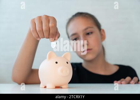 Petite fille avec tirelire à la maison. Une fille triste met une pièce dans la tirelire. Banque D'Images