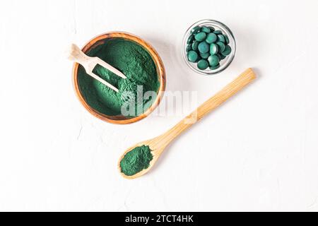 Bol en bois, cuillère et pelle remplis de poudre d'algues spiruline verte sur fond blanc. Un pot en verre avec des comprimés de spiruline. Vue de dessus. Detox. Banque D'Images